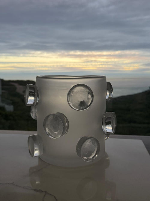 A frosted glass candle holder with clear glass bubble accents, set on a reflective surface with a blurred outdoor coastal landscape in the background.