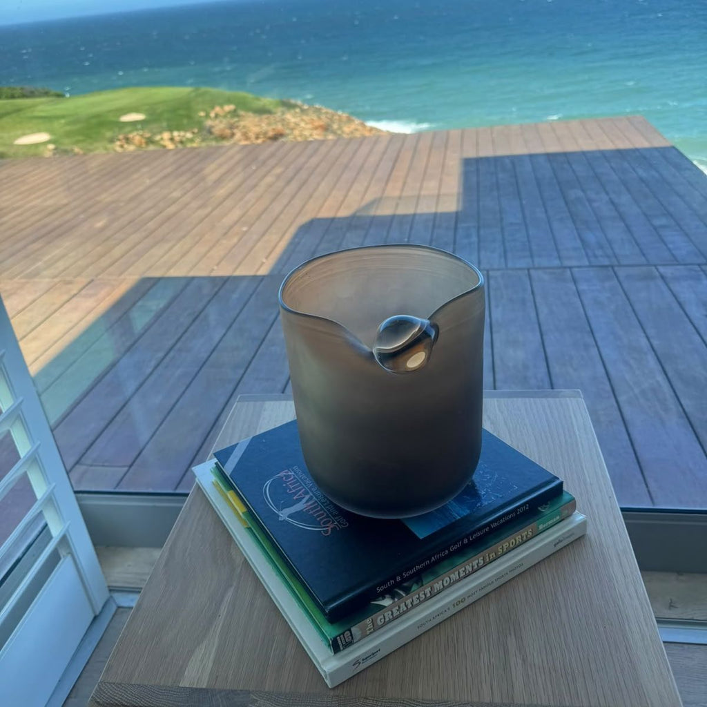 Pebble Dawn Vase on a stack of books on a side table with an ocean-view deck behind — rounded brown glass vase with a frosted satin finish, HMT Home