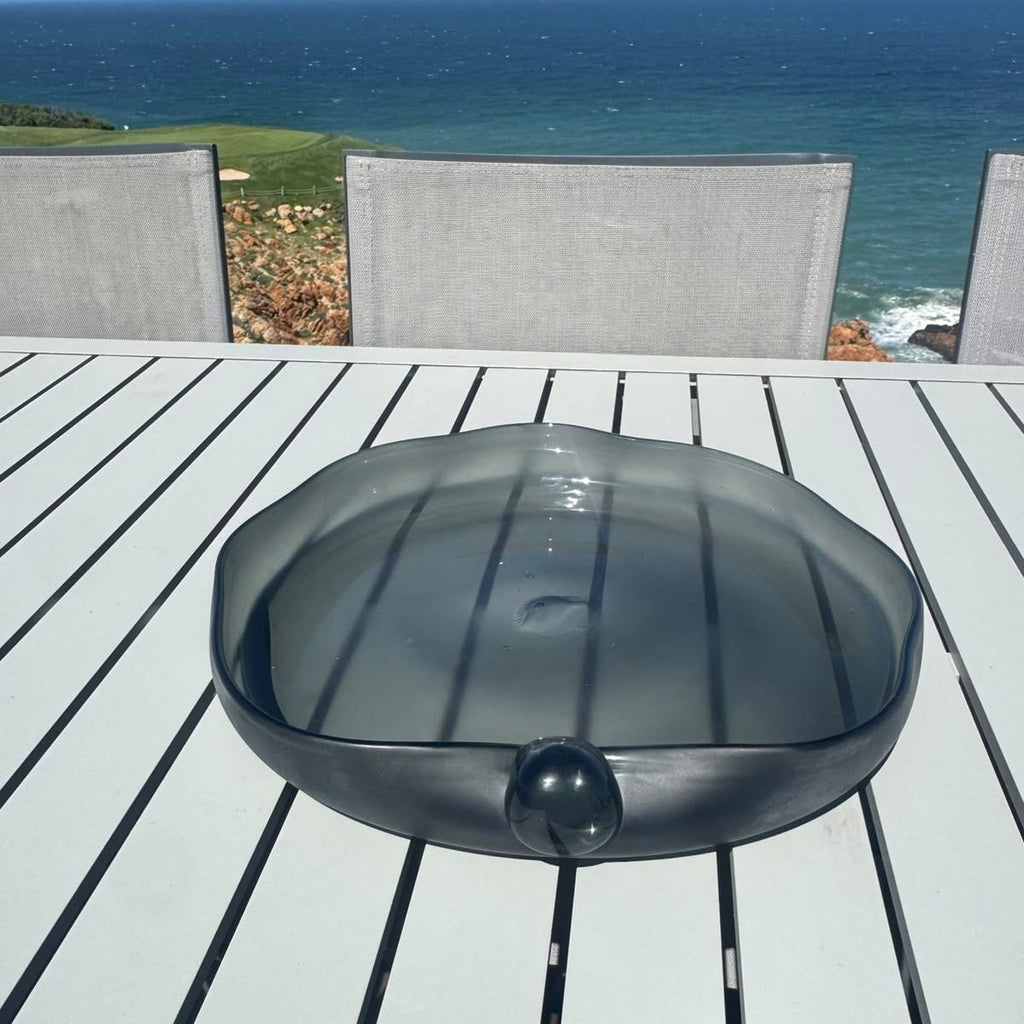 Pebble Eclipse Bowl on a white slatted outdoor table with an ocean view — low-profile organic smoked glass piece, HMT Home