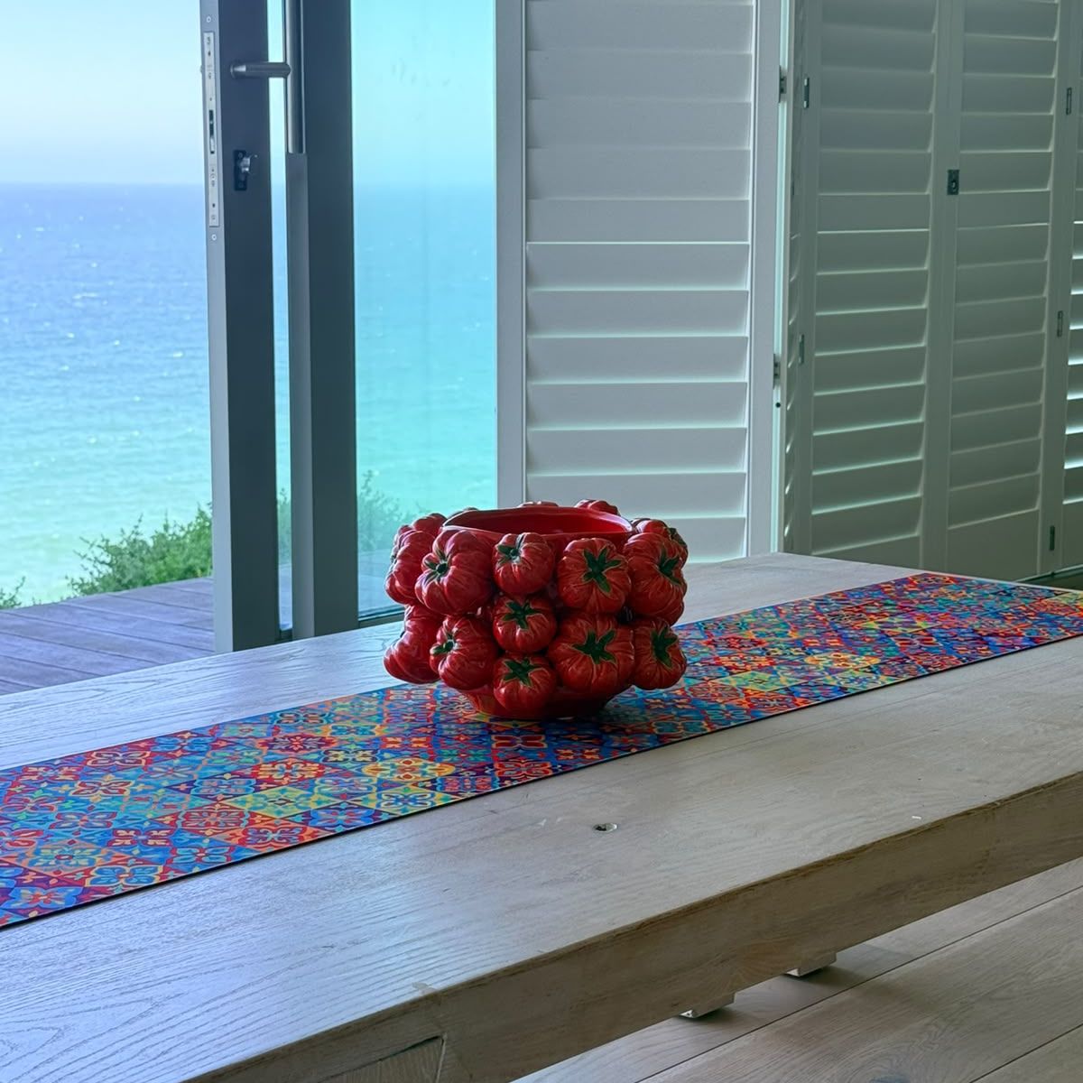 Rosa Tomato Pot on a colourful table runner on a wooden dining table with an ocean view — glossy red ceramic pot covered in sculpted tomatoes, HMT Home