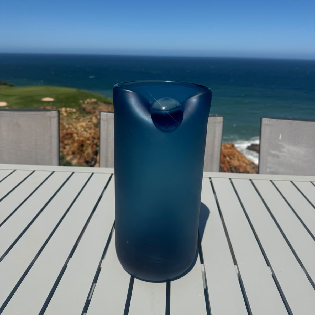 Pebble Tide Vase on a white slatted outdoor table with an ocean view — tall blue glass vase with a satin finish, HMT Home
